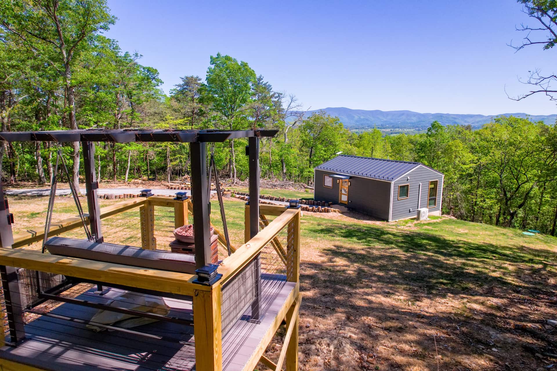 The Turtle's observation deck: a cushioned swinging bench under a steel pergola beside a fire bowl, looking out past the cabin to the valley and the Blue Ridge