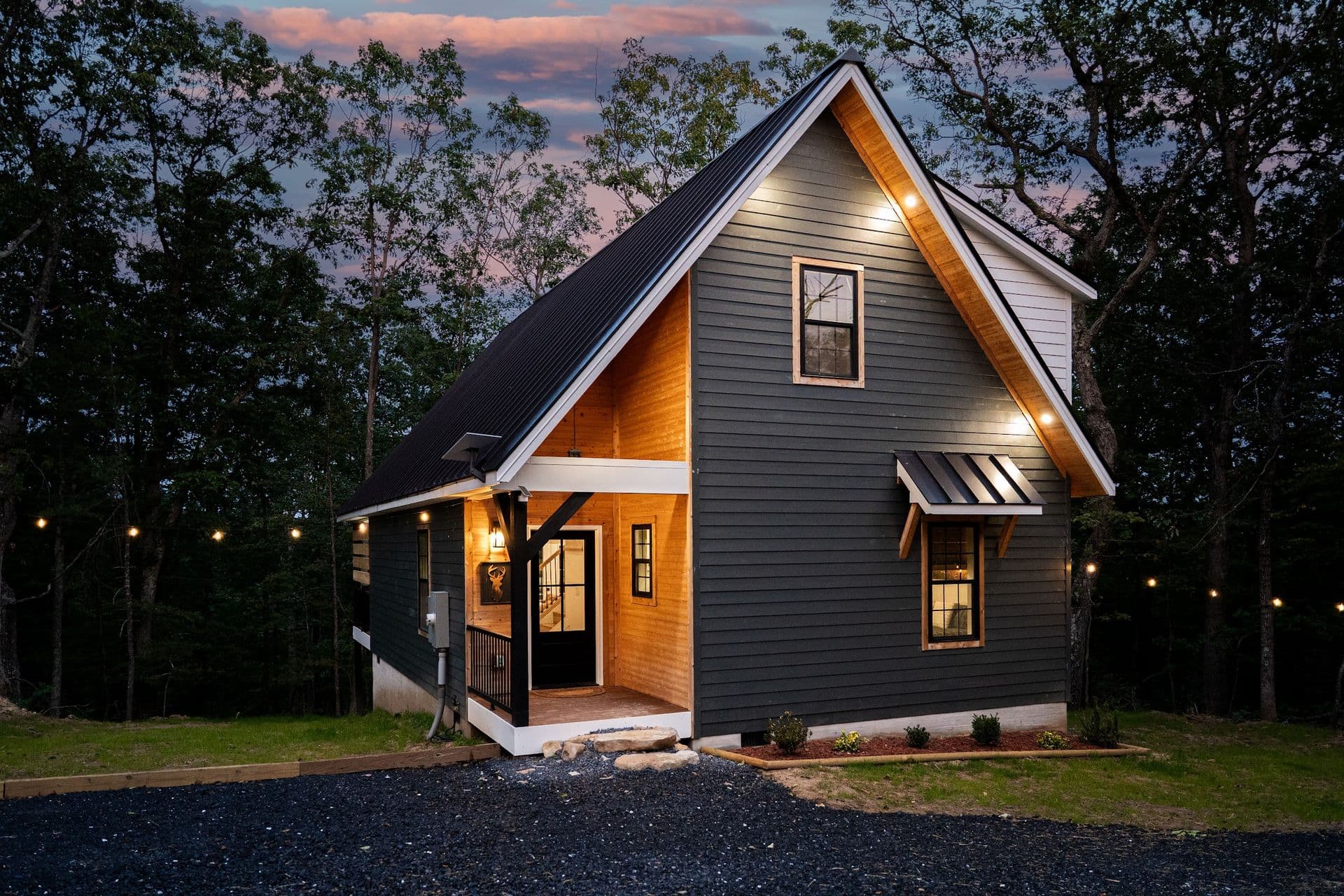 The Stag at dusk: a charcoal cabin with a steep black metal roof and a cedar-lined gable, porch lights on and string lights in the trees
